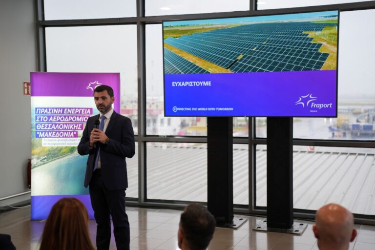 Businessman in a suit speaks into a microphone at a conference, with a large screen showing solar panels behind him and a purple banner to his side promoting green energy in Greece