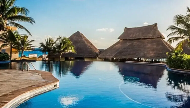 Resort-style infinity pool with thatched huts and palm trees under a clear blue sky.