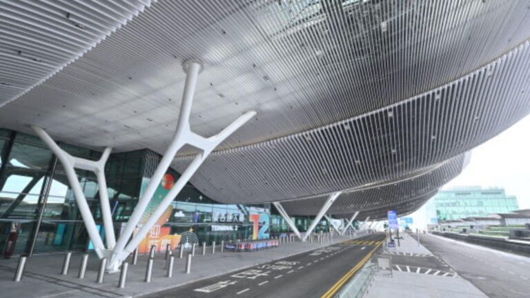 Exterior view of a futuristic building with a large curved metal lattice roof supported by white Y-shaped columns, beside a road with buses and pedestrians edge.