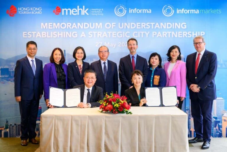 Two people sign documents at a table while ten colleagues stand behind them during a memorandum of understanding ceremony, with a floral centerpiece on the table.