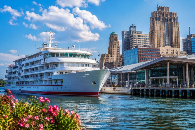 White cruise ship docked at a waterfront terminal with a city skyline in the background on a sunny day.