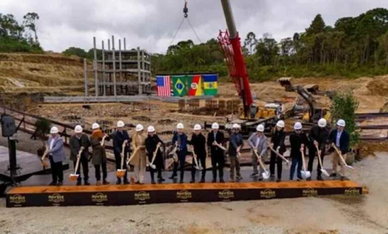 Group of dignitaries in hard hats shoveling earth at a construction site during a groundbreaking ceremony, flags visible in the background