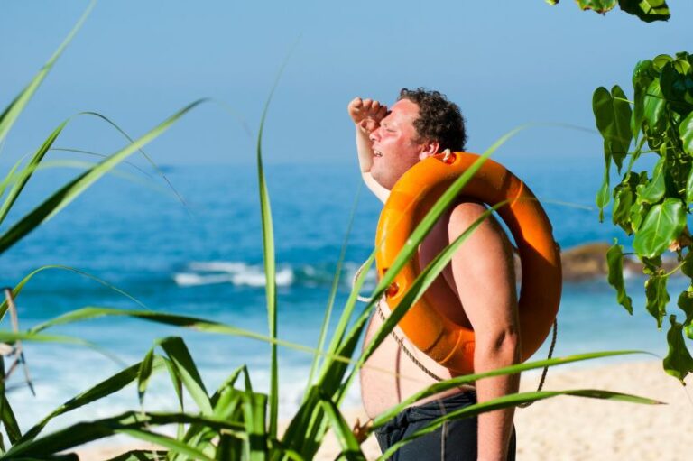 Man on a sunny beach with an orange life preserver around his torso, shielding his eyes as he gazes at the ocean.