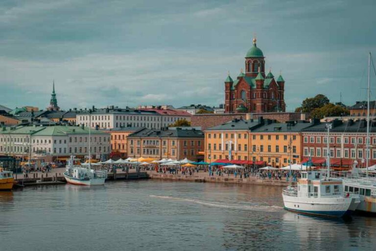 Harbor town with pastel waterfront buildings, a prominent red-brick church with green domes, and boats docked along a calm waterfront promenade.
