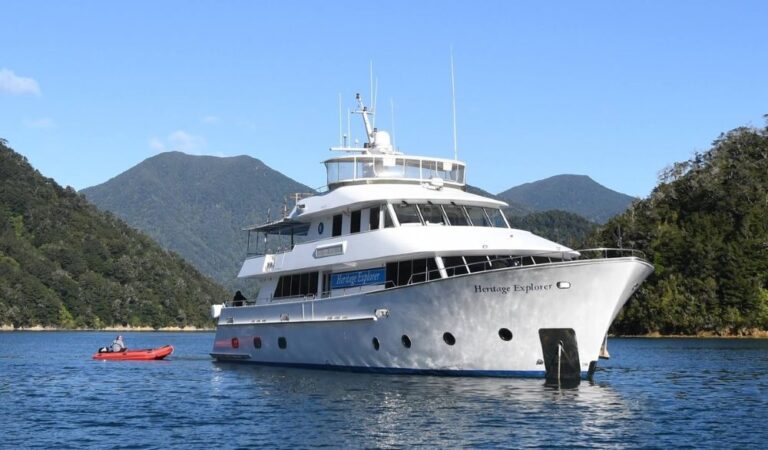 White multi-deck motor yacht named Heritage Explorer anchored in calm blue water near a forested, mountainous coastline.