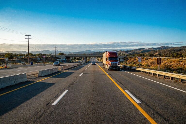 Red semi-truck driving in the right lane of a divided highway on a sunny desert day, with barriers and an overpass in the distance.