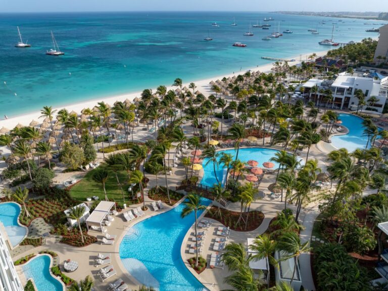 Aerial view of a tropical beach resort with turquoise sea, white sand, palm trees, and curved blue pools surrounded by lounge chairs.