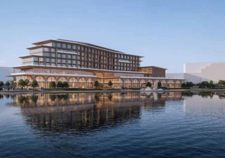 Modern waterfront complex with a stacked glass-and-concrete building, arched ground-floor colonnade, trees, and a calm reflecting pool.