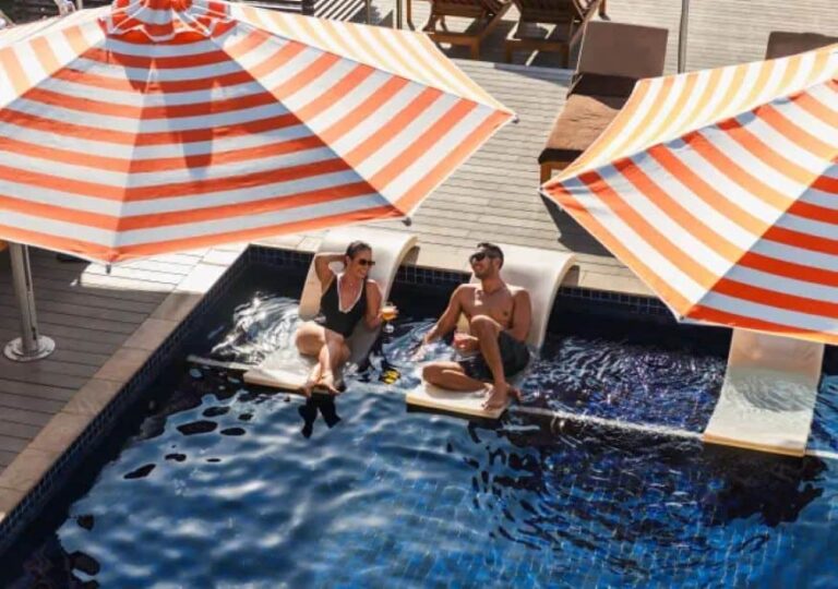 Two people lounge on poolside floating chairs in a blue pool beneath orange-and-white striped umbrellas.