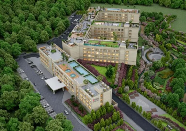 Aerial view of a beige multi-building resort complex with rooftop pools, terraces, and surrounding green forested area.