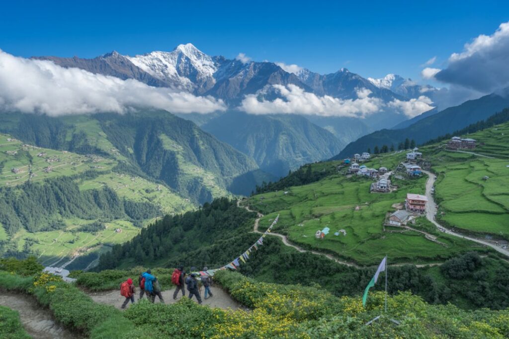 Hikers with red and blue backpacks walk along a dirt path through a green valley with distant snow-capped mountains and a hillside village, under a blue sky.