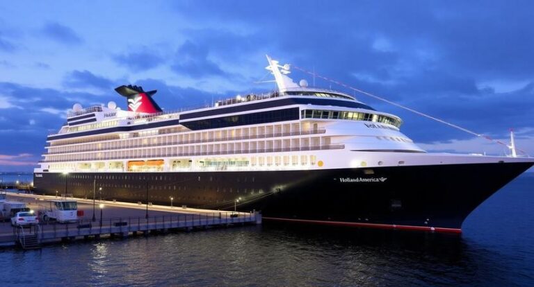Holland America cruise ship docked at a pier at dusk, lights reflecting on calm water.