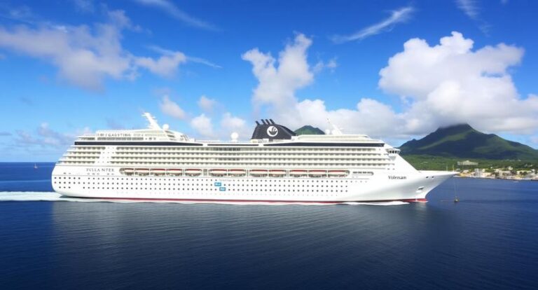 A large white cruise ship with many decks sailing on calm blue water near a green, mountainous coastline under a bright blue sky with scattered clouds.