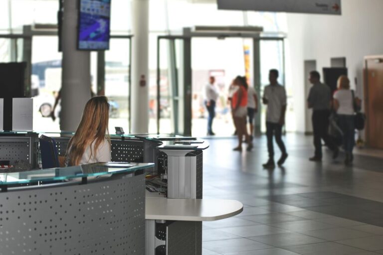 Reception desk with a woman working on a computer in a bright modern lobby; glass doors and people walking in the background.