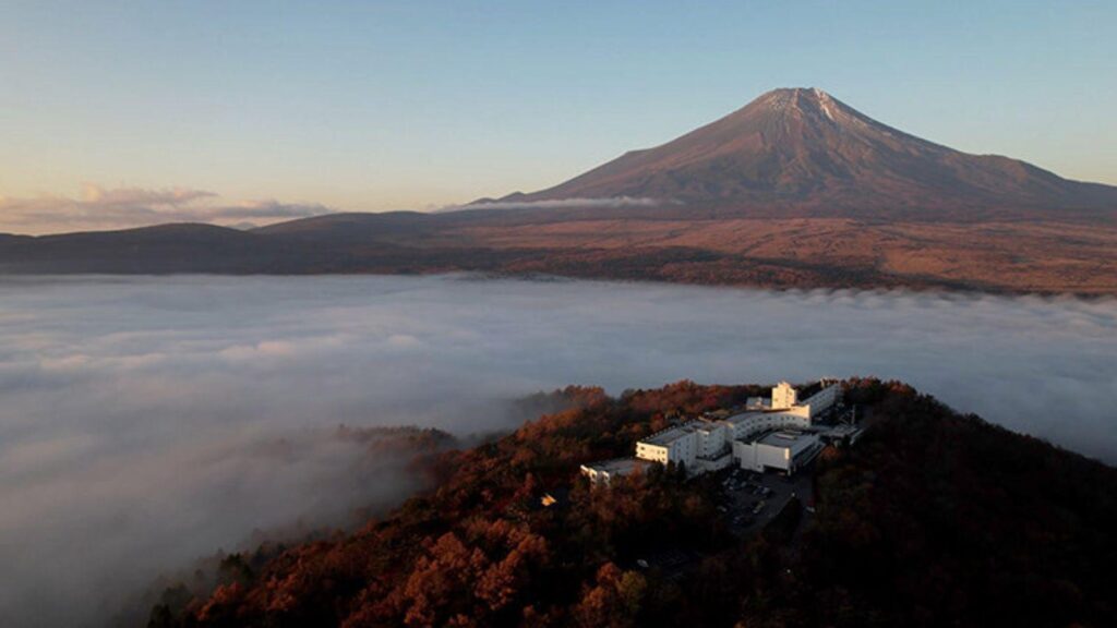 Snow-capped mountain rising above a sea of low-lying clouds, with a white building complex on a forested hillside in the foreground at sunrise.