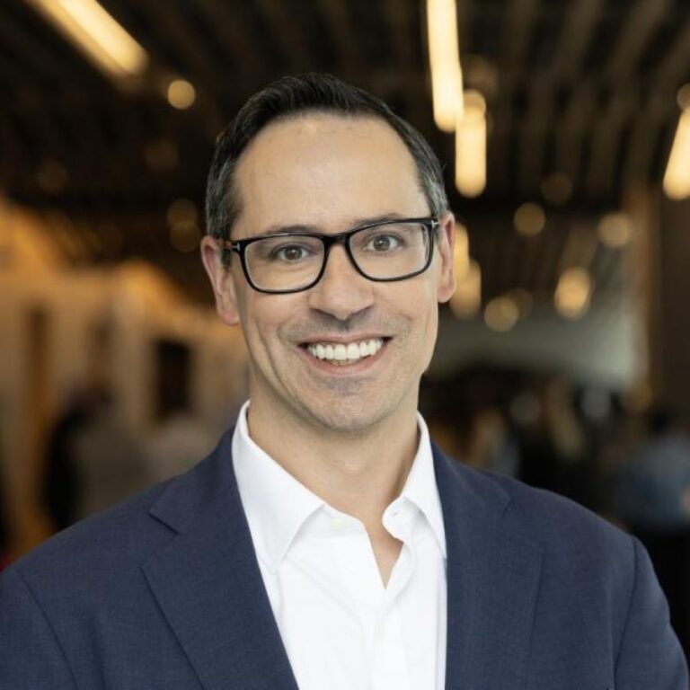 Smiling man wearing glasses and a navy suit jacket in a blurred indoor setting.