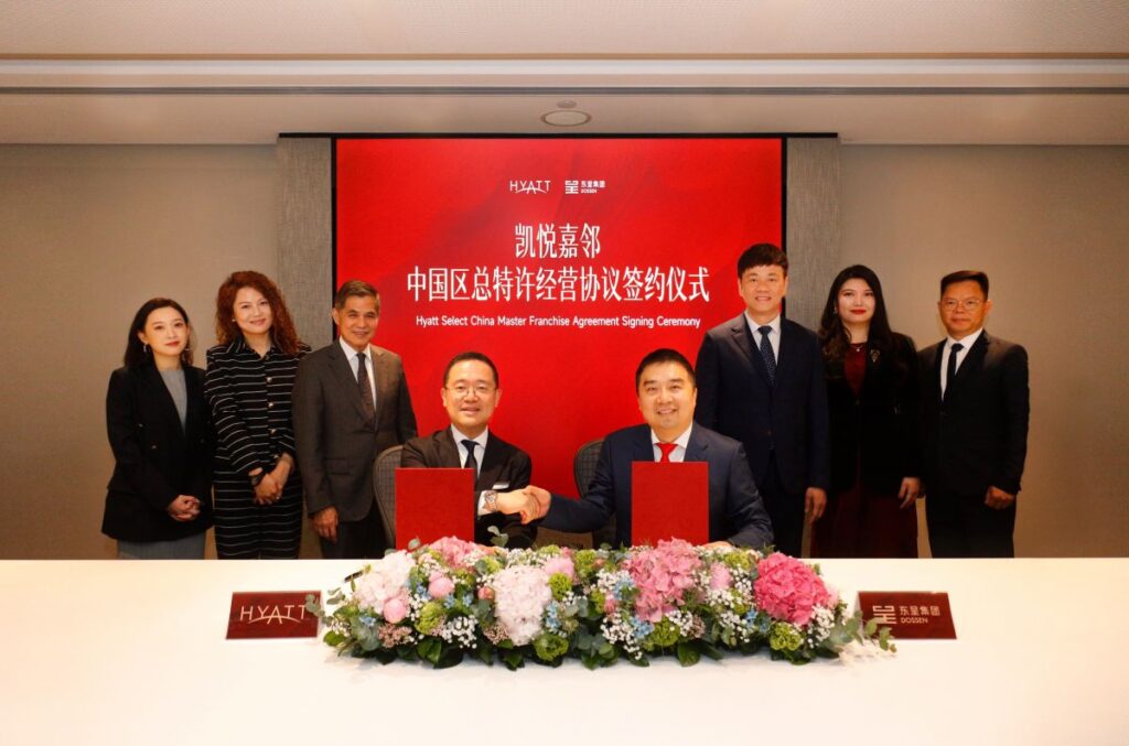 Two men in suits sign red folders at a ceremonial table, flanked by colleagues, with a red backdrop reading Hyatt Select China Master Franchise Agreement Signing Ceremony and blooming floral centerpiece in front.