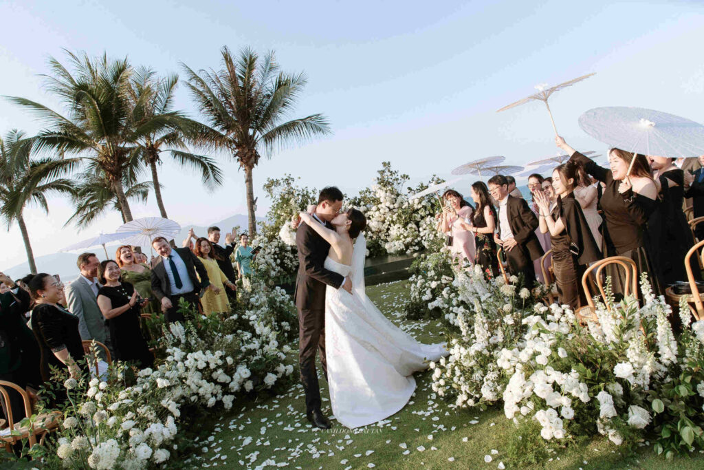 Bride and groom kiss as cheering guests stand along a flower-lined aisle at an outdoor beach wedding with palm trees in the background.