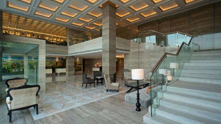 Spacious hotel lobby with marble floors, glass-railed staircase, and beige seating areas.
