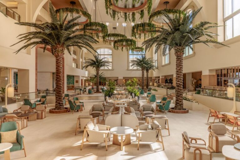 Double-height hotel lobby with tall palm trees, cream seating, and large windows filling the space with natural light.