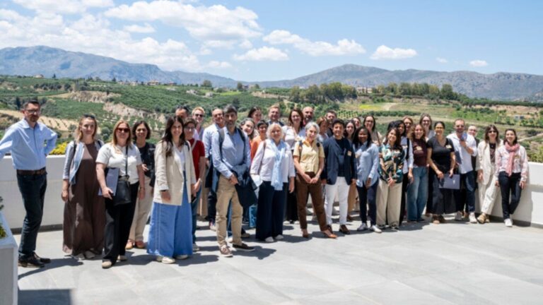 Group of diverse adults posing on a terrace with a rural hillside landscape and mountains in the background, sunny day.
