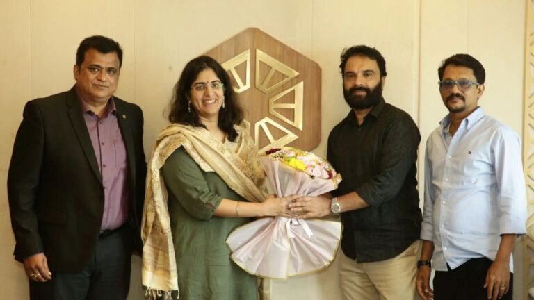 Four professionals stand together as a woman in green accepts a bouquet from a bearded man, in front of a wooden geometric logo on the wall.