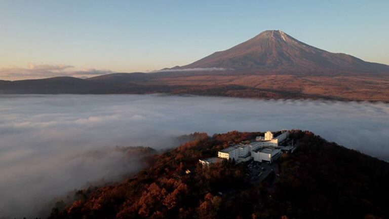 Mountain peak rising above a sea of clouds, with white buildings on a forested autumn hillside below.