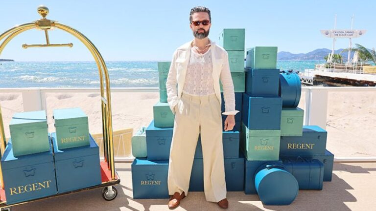 Man with a beard in a light cream suit and sunglasses stands beside stacked Regent boxes on a sunny beach by the sea.
