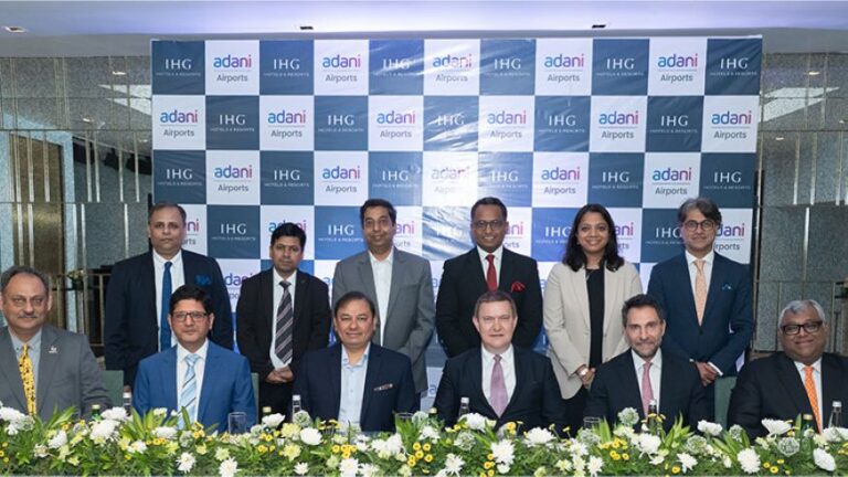 Group of business professionals posing for a photo at a corporate event, backdrop covered with adani Airports and IHG logos; floral centerpiece on the table.