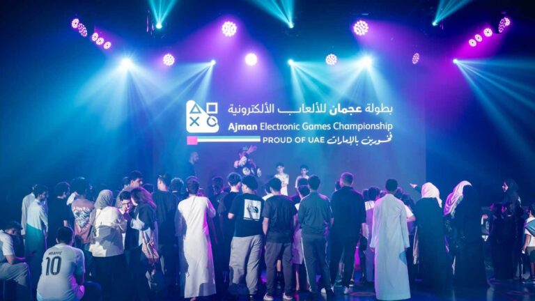Crowd of people gathered in front of a stage with colorful lights and a large screen reading 'Ajman Electronic Games Championship' in Arabic and English.