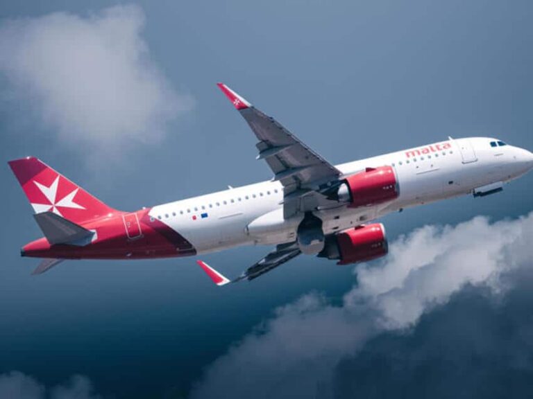 Airplane with Malta airline livery ascending through a cloudy blue sky.