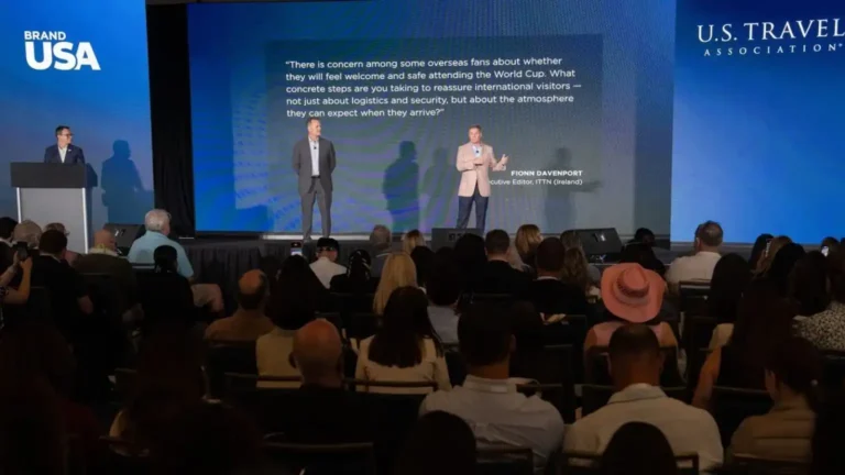 Two men speak on a stage at a conference; a large screen behind them displays a quote and their names, with audience seated in front and speaker podium on the left.