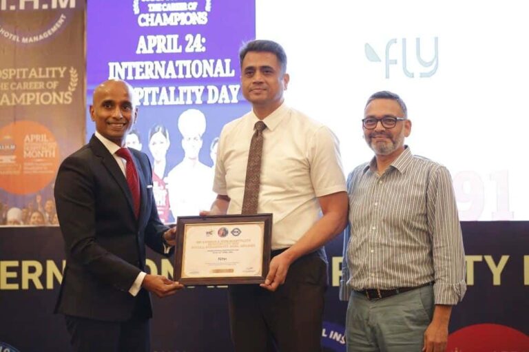 Three men on stage pose with a framed certificate during an awards ceremony, two in business attire and one in a striped shirt, backdrop shows event banners.