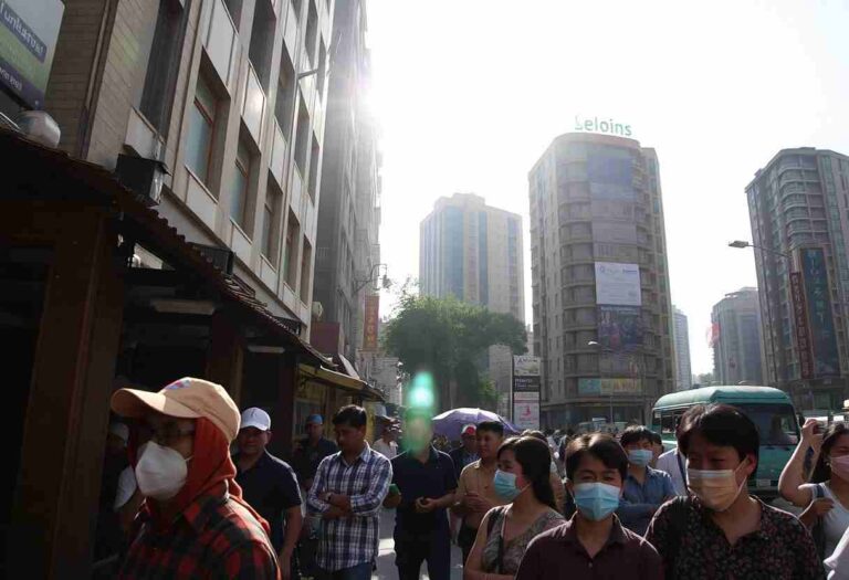 Crowded urban street scene with pedestrians wearing masks, sunlight glare from tall buildings, and a teal bus in the background.