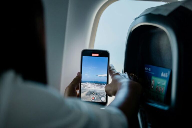 Passenger holds up a smartphone to capture the airplane window view, showing blue sky and clouds on the screen while seated by the window