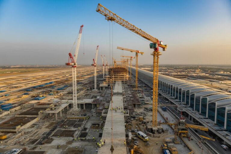 Tall construction cranes tower over a large building site with workers and concrete platforms, stretching toward a long modern terminal building on the right at dusk.