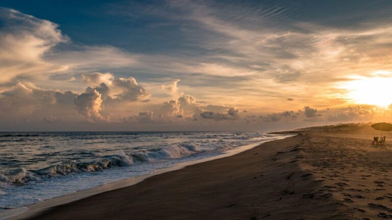Golden sunset over a sandy beach with gentle waves and a small umbrellaed seating area on the right.