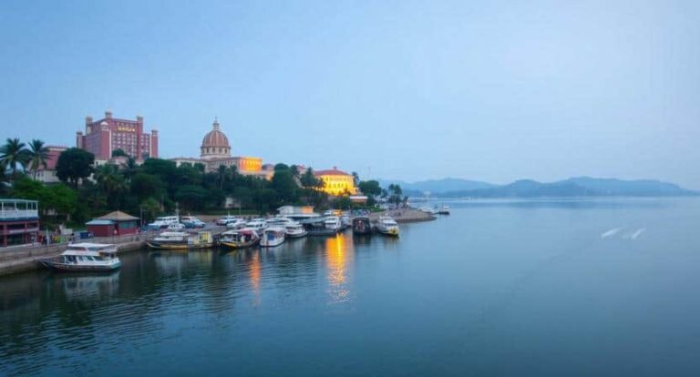 Harbor scene at dusk with boats docked along a waterfront and a domed building on a hillside amid trees.