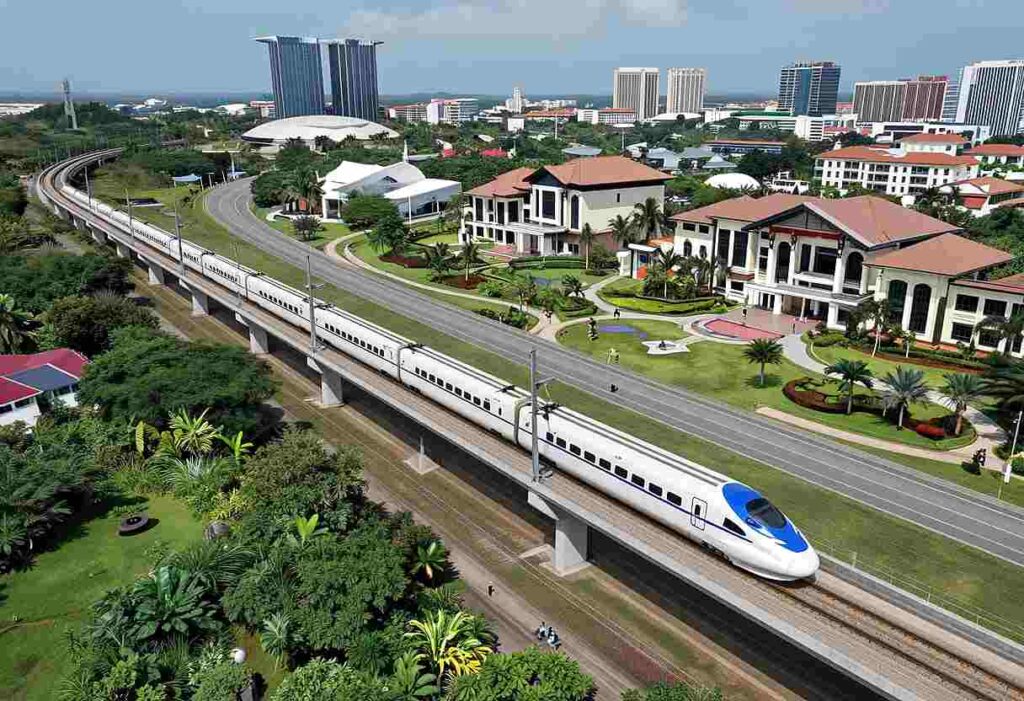 A high-speed white train with a blue nose travels on an elevated rail line through a green, modern cityscape with tall buildings in the background.