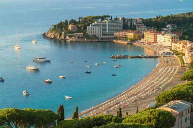 A Mediterranean coastal town with a curved sandy beach lined with umbrellas, calm turquoise water, and pastel buildings along the shore.