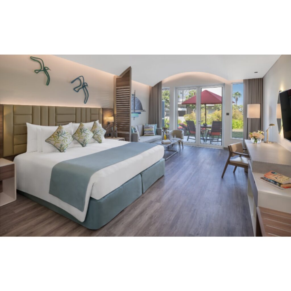 Hotel suite with king bed, teal throw blanket, and a sitting area leading to a garden-view balcony outside the glass doors.