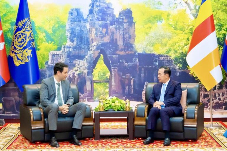 Two men in business suits sit in armchairs across from each other in a formal meeting room with a small table of flowers between them, flags on either side, and a temple ruins backdrop behind them.
