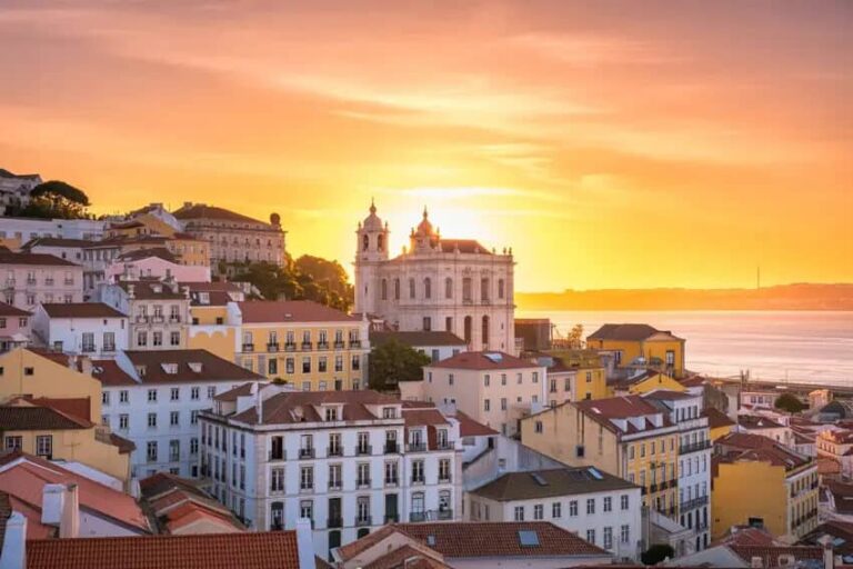 Sunrise over a hilltop city with pastel houses and a prominent church overlooking the water at sunset