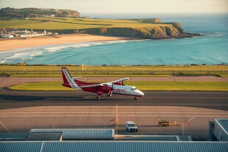 Small red-and-white passenger airplane taxiing on a runway by the coast, with a sandy beach, green fields, and blue sea in the background.