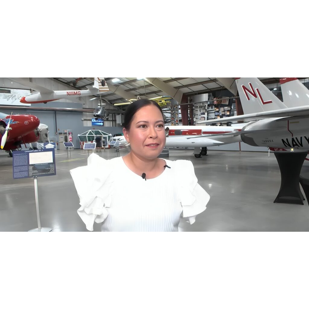 Woman in white blouse being interviewed inside an airplane museum with vintage planes in the background.