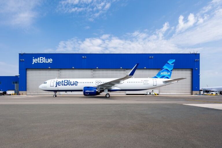 JetBlue airplane on the tarmac in front of a large blue JetBlue hangar with branding visible on the building and tail fin.