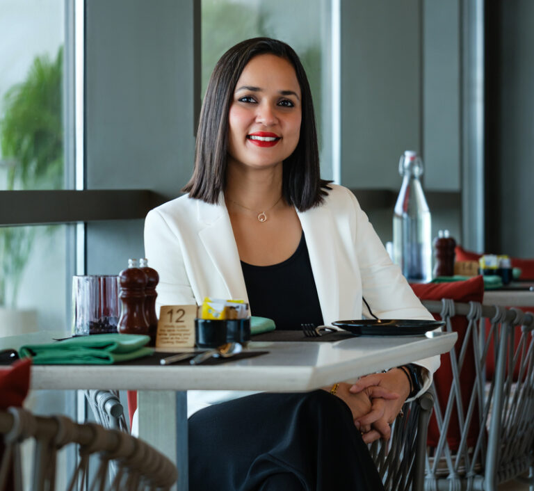 Woman in a white blazer sits at a cafe table, smiling at the camera with a black top underneath.
