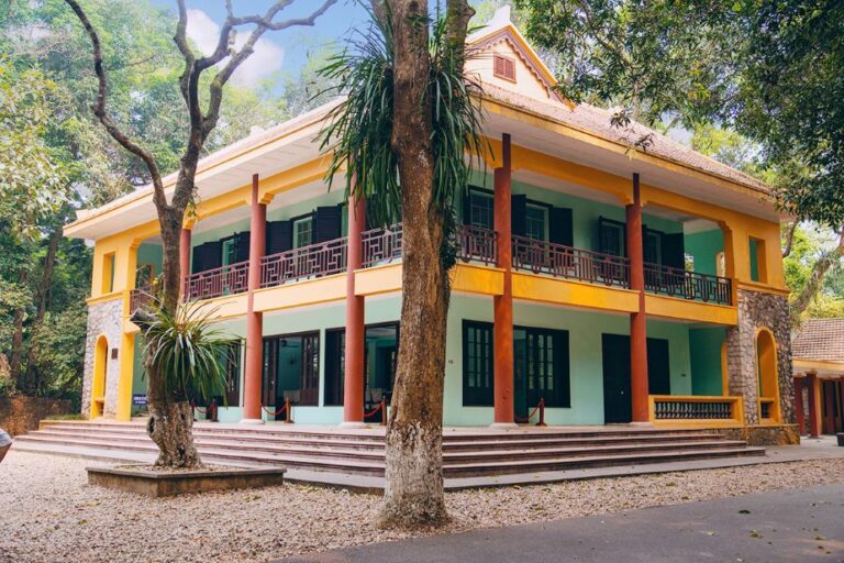 Two-story colorful colonial-style building with yellow and teal walls, red columns, and surrounding trees at the campus.