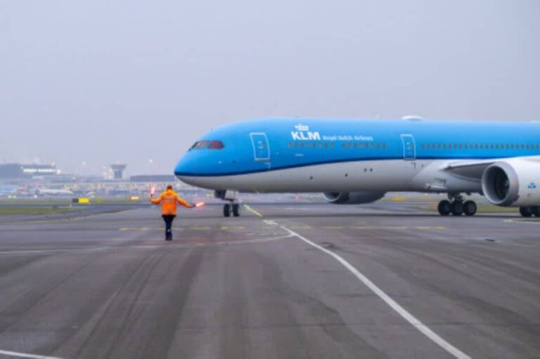 Ground crew member in an orange jacket guides a blue KLM airplane on the tarmac.