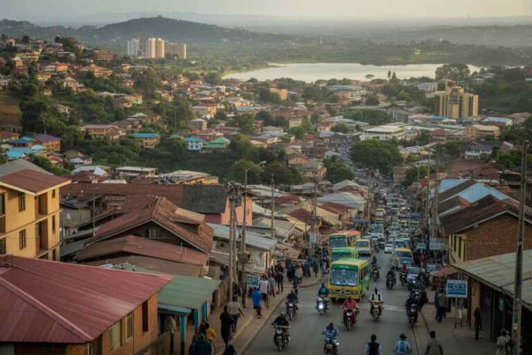 Bustling hillside town street with cars, motorcycles, and pedestrians; colorful rooftops and low houses on both sides; a green bus in the center and a river with hills in the distance.